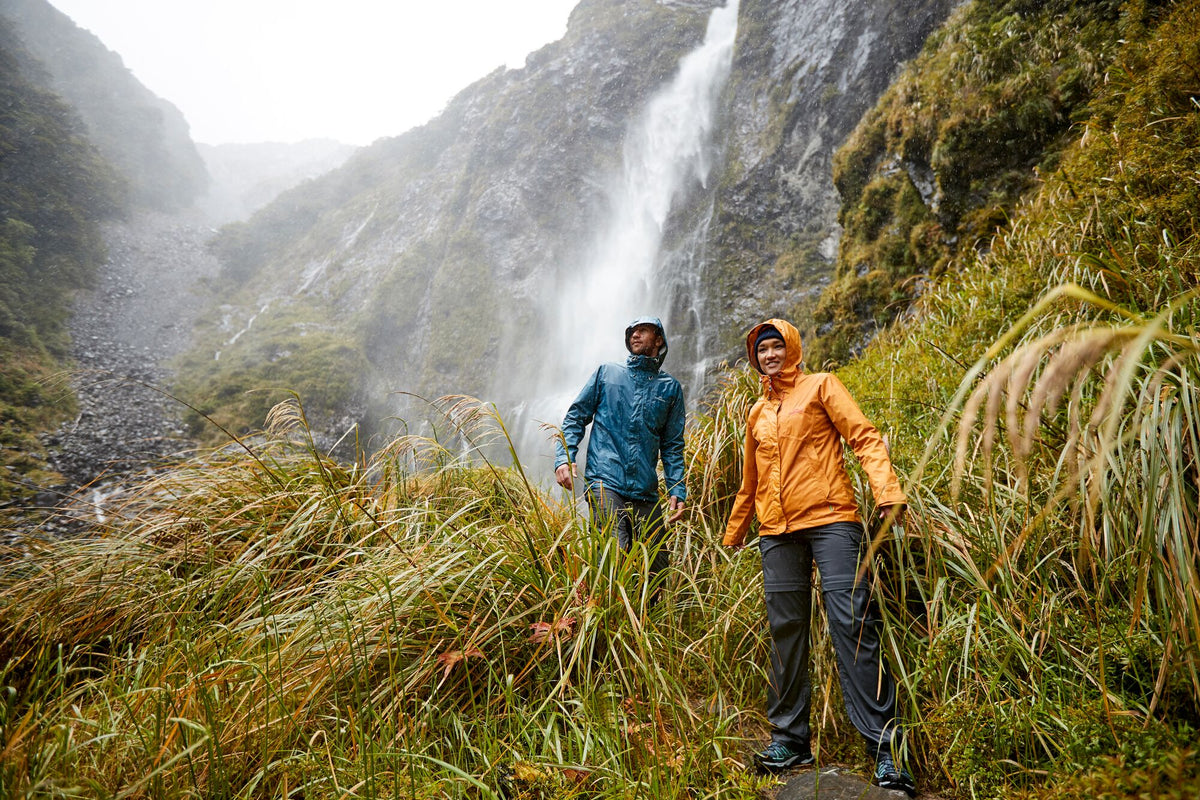 A couple walking through wet long grass in front of a waterfall.