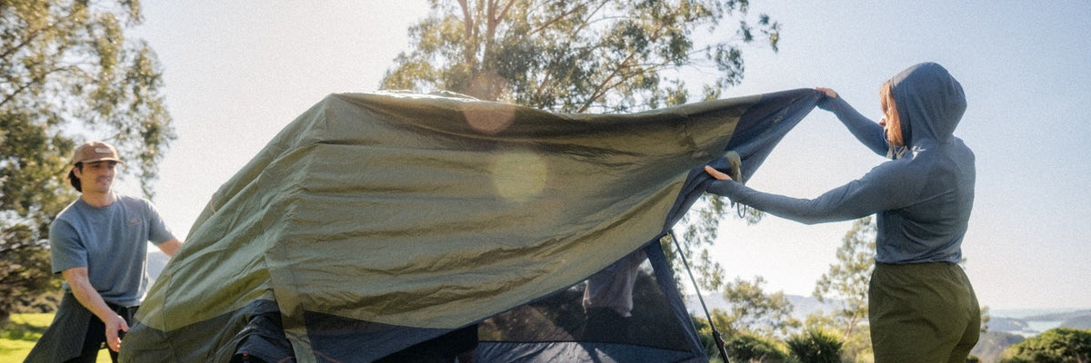 Friends are setting up a tent outdoor, pulling a large green rainfly over the tent frame in a sunny park-like setting with trees in the background. The scene highlights camping preparation and outdoor adventure equipment.
